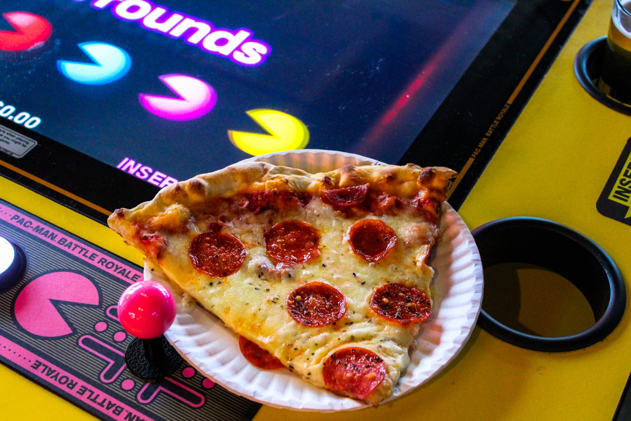 A slice of pepperoni pizza on a paper plate, next to an arcade game featuring colorful Pac-Man graphics and a joystick.