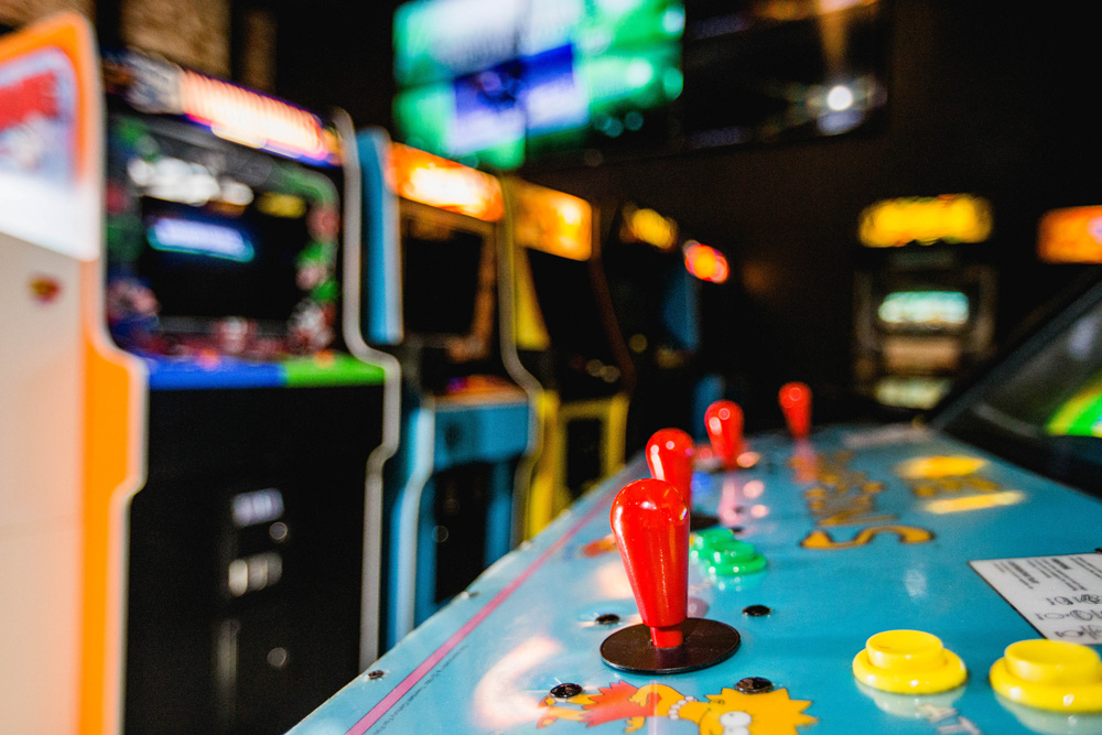 Close-up of a vintage arcade game control panel, featuring colorful joysticks and buttons, with arcade cabinets softly blurred in the background.