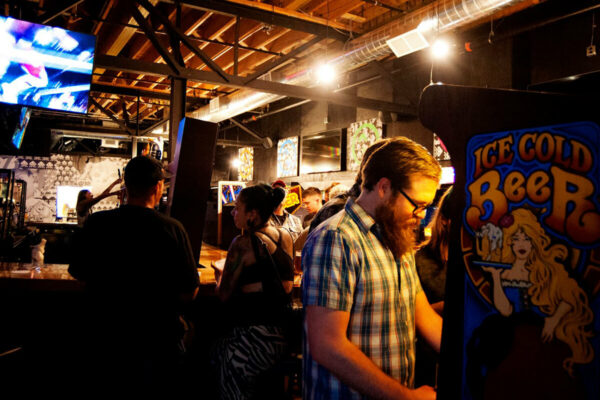 A low-lit interior shot of a bar, showing a bearded man in a plaid shirt playing an arcade game. A low-lit interior shot of a bar, showing a bearded man in a plaid shirt playing an arcade game.
