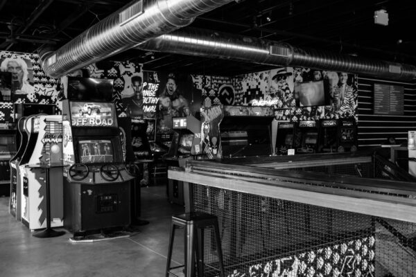 Greyscale shot of a bar's interior with arcade games on the left and a long bar with stools on the right Greyscale shot of a bar's interior with arcade games on the left and a long bar with stools on the right