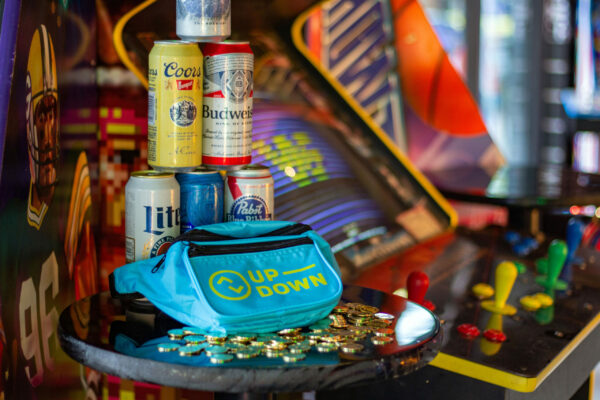 A shot of a teal fanny pack on a pile of gold coins, with stacks of beer cans and an arcade machine in the background A shot of a teal fanny pack on a pile of gold coins, with stacks of beer cans and an arcade machine in the background