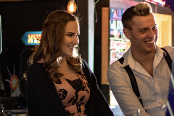 A medium shot of a smiling woman with long brown hair, wearing a printed dress and a cardigan, next to a man. A medium shot of a smiling woman with long brown hair, wearing a printed dress and a cardigan, next to a man.