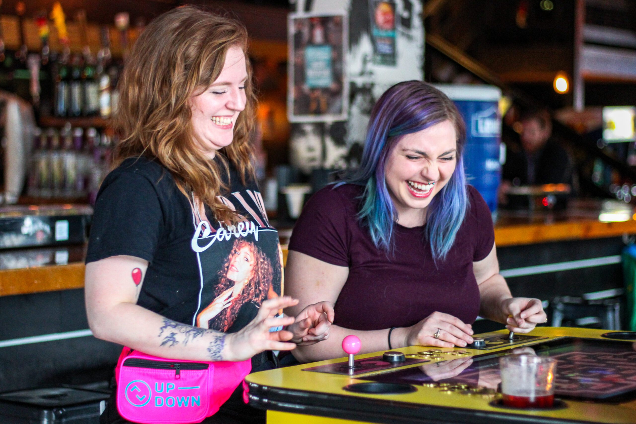 Two women enjoying a game of pinball at a lively bar, focused on the machine and having fun together