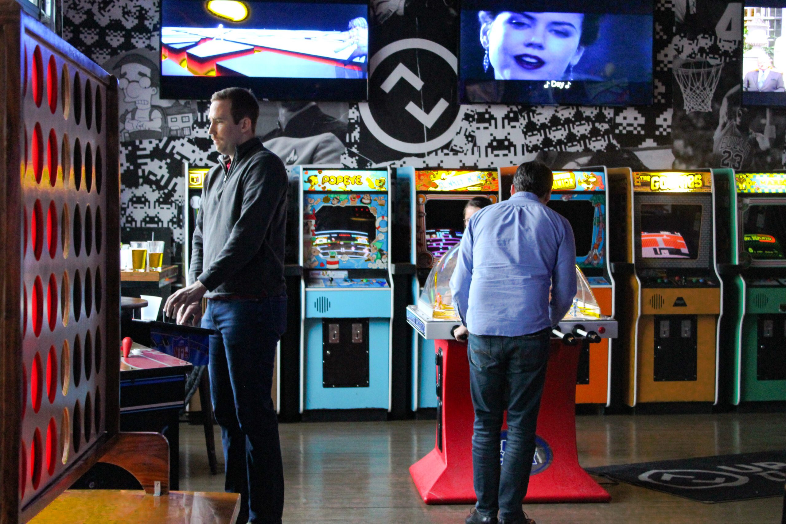 Two men stand in front of colorful arcade machines, engaged in a gaming session at a lively arcade