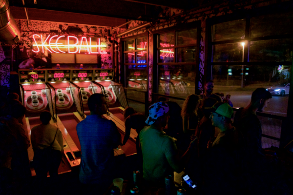 Several friends stand around a vibrant pinball machine, engaged in play and sharing laughter in a lively arcade setting Several friends stand around a vibrant pinball machine, engaged in play and sharing laughter in a lively arcade setting