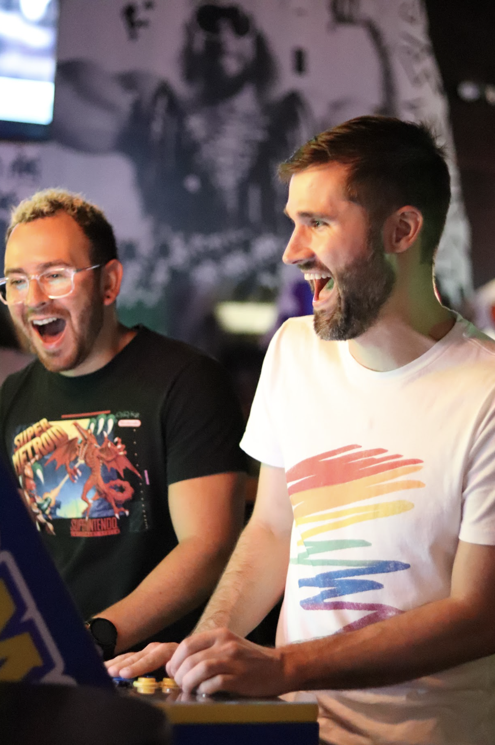 A medium shot of two smiling men, one wearing glasses and a dragon t-shirt and the other a rainbow t-shirt.