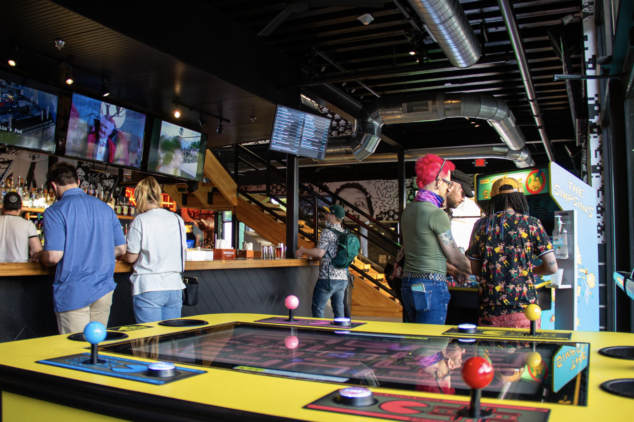 A low-angle interior shot of a bar, showing a yellow Pac-Man table, a bar counter, and people mingling