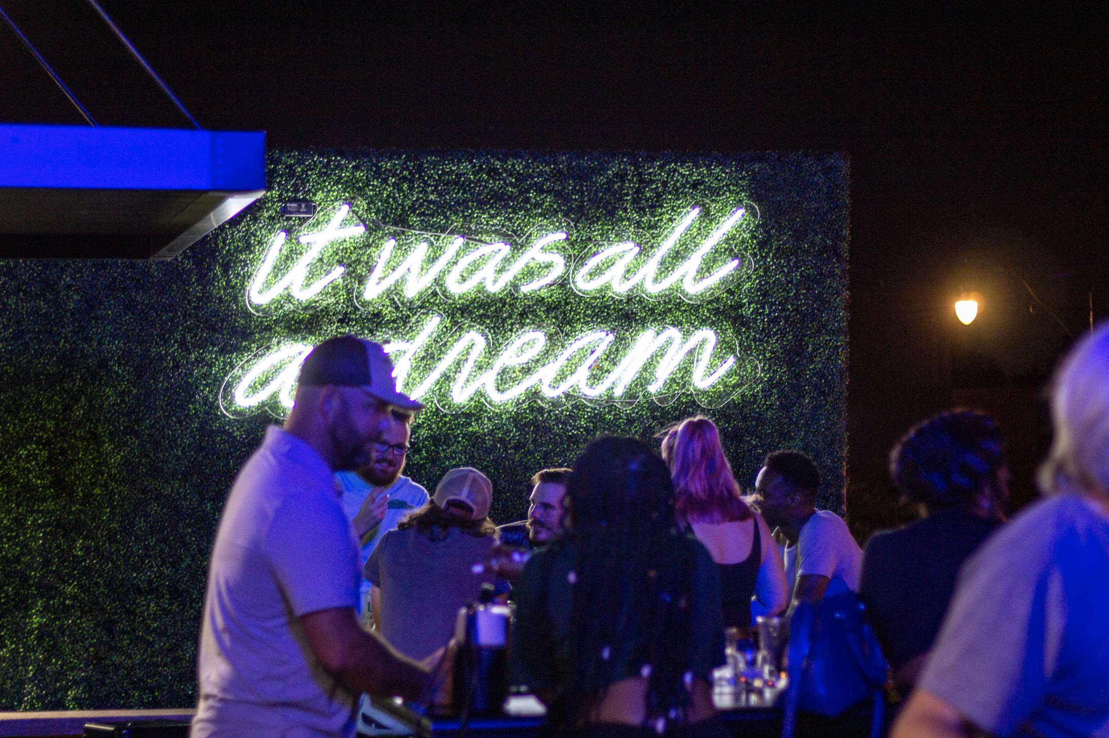 A night shot of a bar patio with people gathered in front of a neon sign that reads "it was all a dream.