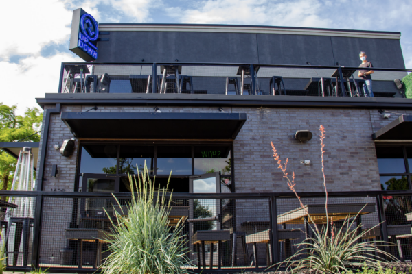 A daytime exterior shot of a bar with a black brick facade, a deck with tables, and a blue neon sign. A daytime exterior shot of a bar with a black brick facade, a deck with tables, and a blue neon sign.