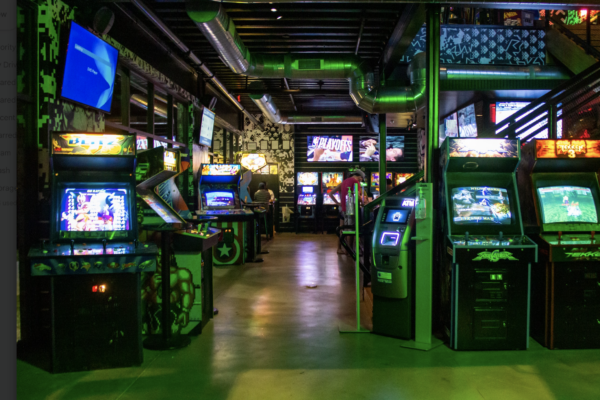 A wide, low-light shot of a bar's interior, showing a long hallway lined with illuminated arcade games. A wide, low-light shot of a bar's interior, showing a long hallway lined with illuminated arcade games.