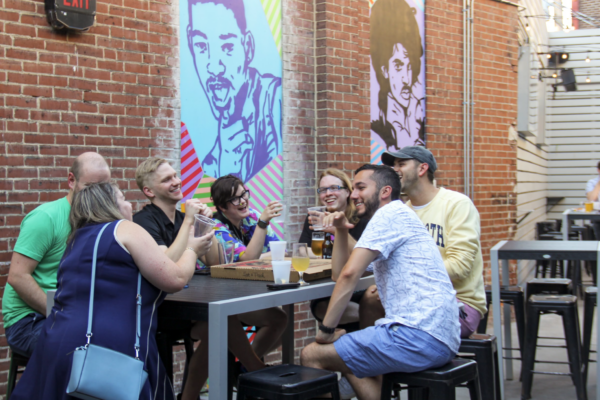 A wide shot of a group of friends laughing and drinking at a table on a patio with murals on a brick wall. A wide shot of a group of friends laughing and drinking at a table on a patio with murals on a brick wall.