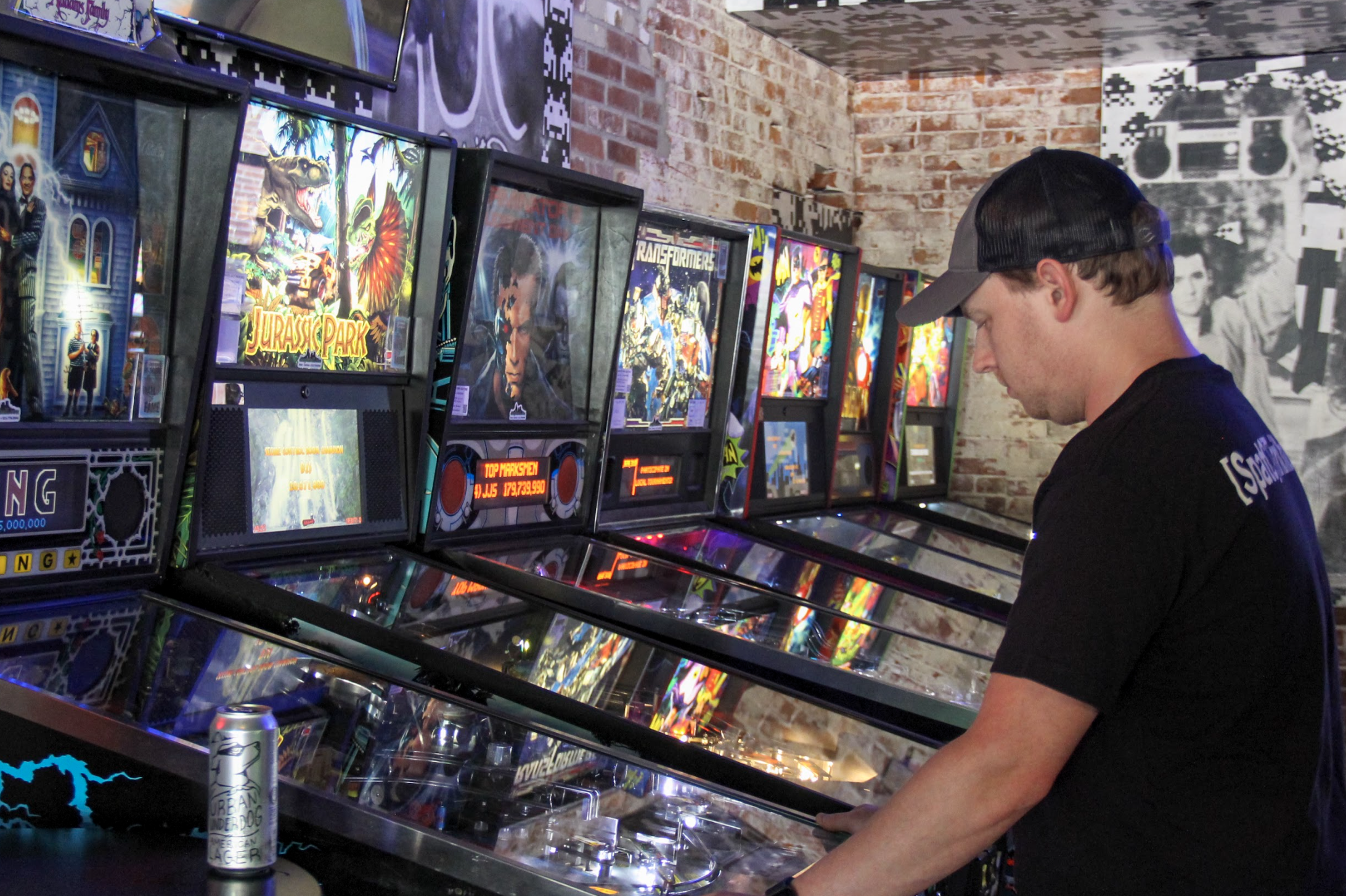 A medium shot of a man in a black shirt and cap playing a "Transformers" pinball machine.