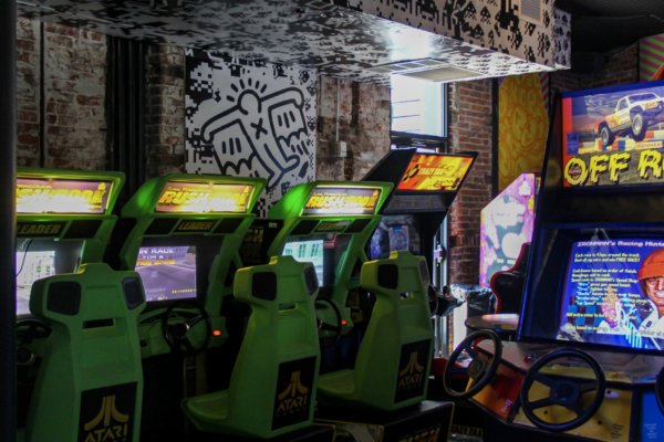 A row of green racing arcade games with seats and steering wheels in a room with graffiti-style murals. A row of green racing arcade games with seats and steering wheels in a room with graffiti-style murals.