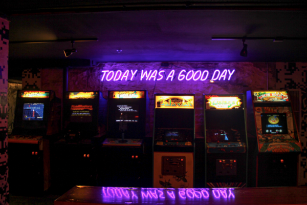 A wide, low-light shot of a row of arcade machines under a purple neon sign that reads, "Today Was A Good Day." A wide, low-light shot of a row of arcade machines under a purple neon sign that reads, "Today Was A Good Day."