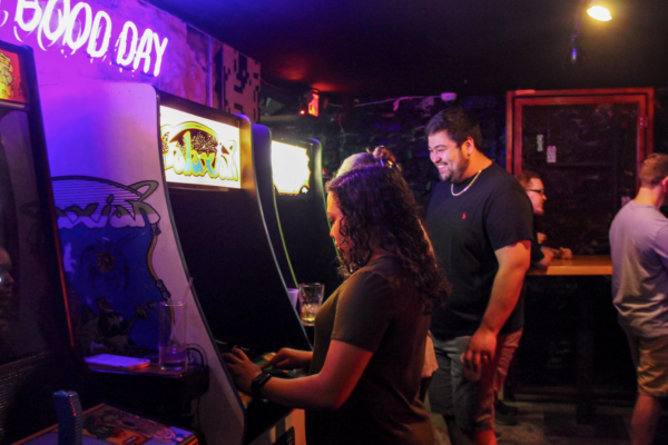 A low-light shot of a woman playing an arcade game as a man stands behind her smiling. A low-light shot of a woman playing an arcade game as a man stands behind her smiling.