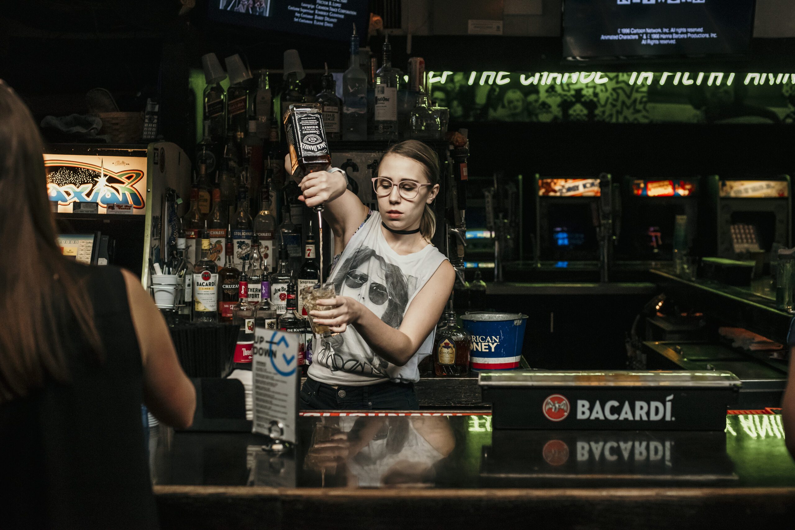 A woman pours a drink from a bottle into a glass at a bar, surrounded by various bottles and a lively atmosphere