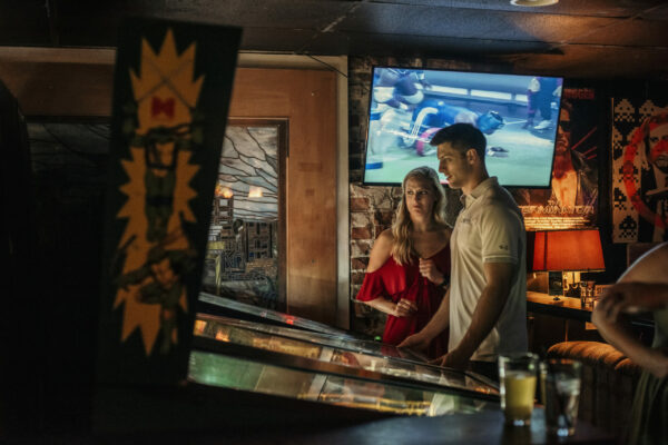 A man and woman pose happily in front of a vintage pinball machine, surrounded by bright lights and arcade games. A man and woman pose happily in front of a vintage pinball machine, surrounded by bright lights and arcade games.