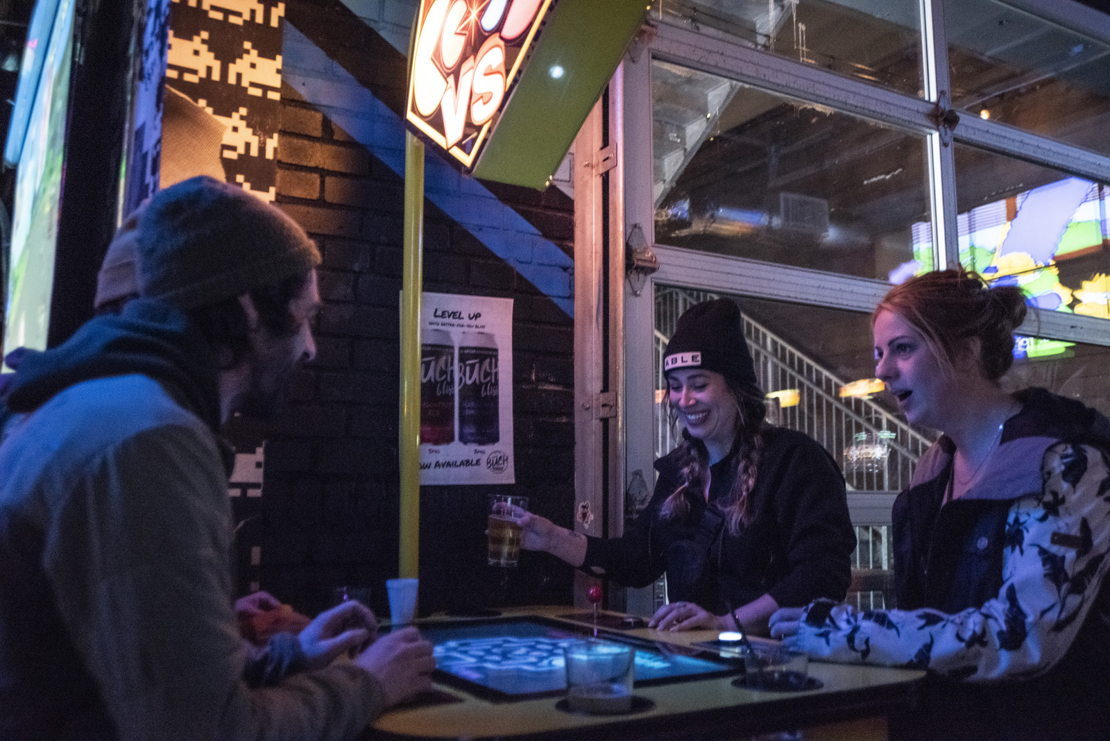 A group of three friends seated at a table, each with a beer, engaged in lively conversation